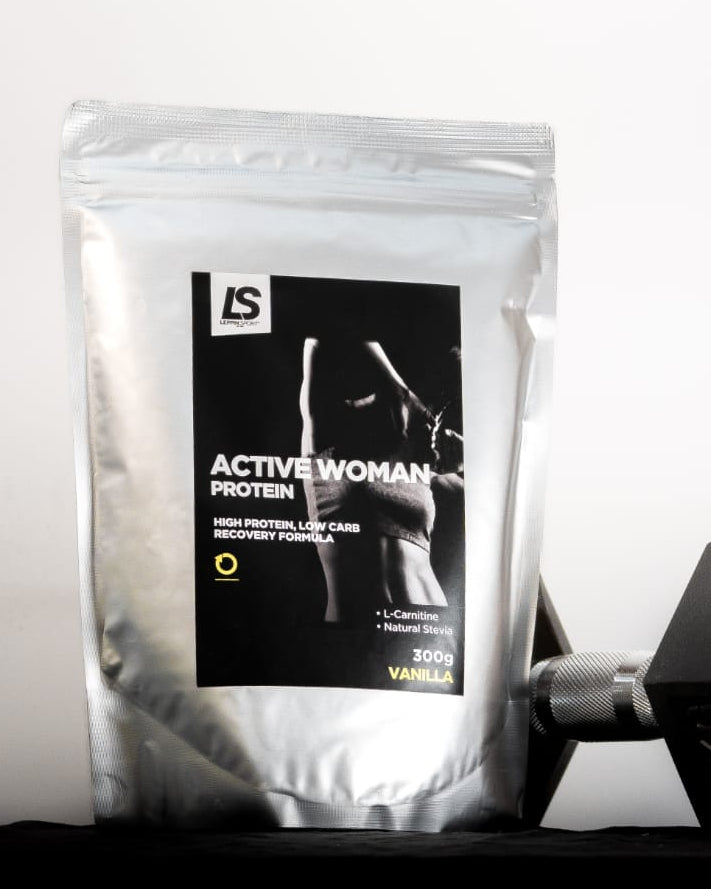 A silver stand-up pouch of IS Active Woman Protein powder with a black label, placed next to a black hexagonal dumbbell on a dark surface against a white background.