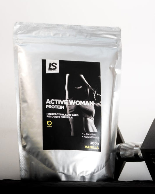 A silver stand-up pouch of IS Active Woman Protein powder with a black label, placed next to a black hexagonal dumbbell on a dark surface against a white background.