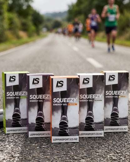 Five boxes of Leppin'sport Squeezezy Energy Gel are lined up on a paved road with blurred runners in the background.